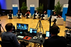 In einem Halbrund sitzen sieben Personen auf weißen Sesseln. Im Vordergrund bedienen zwei Personen die Technik.